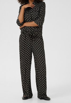 Woman wearing black matching top and wide-leg pants with white circular patterns, standing with arms crossed and hand touching chin.