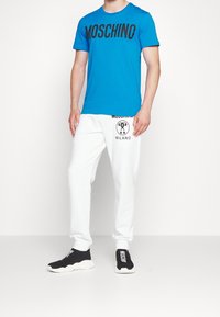 Muž oblečený v jasně modrém tričku Moschino, bílých teplácích Moschino Milano a černých mokasínech Moschino stojící na bílém pozadí.