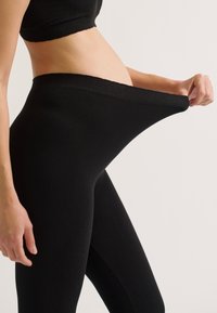 Zwarte geribde leggings met een hoge tailleband, uitgerekt naar de zijkant, wat flexibiliteit en textuur van het materiaal toont. Simpel ontwerp, geen duidelijke patronen.