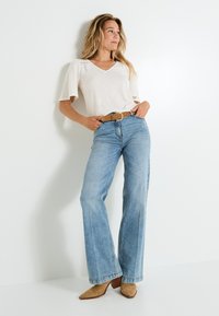 Top de color beige claro con escote en V y mangas cortas, combinado con jeans de campana azul claro y botas de tobillo de tacón color marrón, luciendo un cinturón marrón.