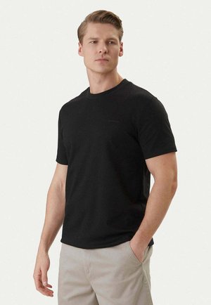 Giovane uomo che indossa una t-shirt nera semplice a maniche corte e pantaloni beige chiaro, in piedi con una mano in tasca davanti a uno sfondo uniforme.