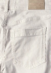 Tejido claro a rayas con textura de mezclilla. Presenta un bolsillo trasero y una etiqueta de cuero rectangular en la cintura con un logo grabado.