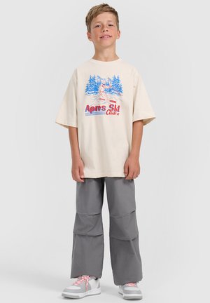 Chłopiec stoi, ubrany w oversize kremowy T-shirt z grafiką narciarza i tekstem, szare luźne spodnie oraz białe trampki z różowymi sznurówkami.