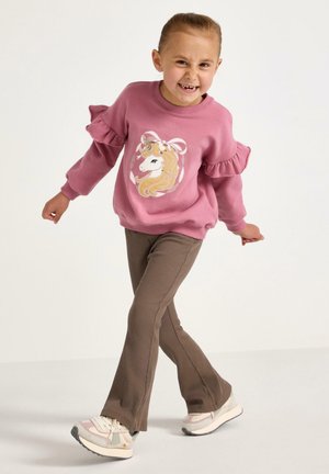 Rosa sweatshirt med volangärmar som har en enhörningsgrafik, tillsammans med bruna ribbade byxor och flerfärgade sneakers.