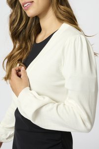 Bílý pletený cardigan s nabíranými rukávy, s výstřihem do V. Nosený přes černý top, zdůrazňující jemnou strukturu a uvolněný střih.