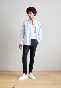 Lichtblauw, gestreept overhemd met knopen, gedragen over een witte tanktop, gecombineerd met zwarte broeken en witte sneakers, op een houten vloer.