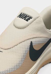 Κοντινό πλάνο της φτέρνας ενός μπεζ αθλητικού παπουτσιού Nike που δείχνει υφή υφάσματος, το μαύρο λογότυπο Nike swoosh και το κείμενο "NIKE" στην ετικέτα της φτέρνας.