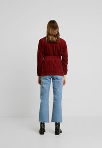 Bordeaux corduroy jas met een strikceintuur, gecombineerd met lichtblauwe flare-jeans en zwarte hoge laarzen. Gestructureerde stof met ribbelpatroon.