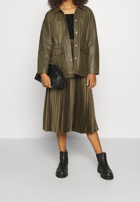 Oliivinvihreä nahkatakki, jossa on kaksi etutaskua, yhdistettyinä pleatattuun, polvipituiseen oliivinvihreään hameeseen ja mustiin nilkkureihin. Malli pitää mustaa clutch-laukkua.