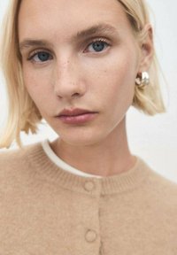 Cárdigan de lana beige con cuello redondo, textura de punto y botones. La modelo tiene el pelo corto rubio y lleva pendientes de aro.