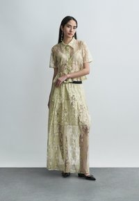 Γυναίκα με διαφανές κίτρινο δαντελωτό μπλουζάκι με κοντά μανίκια και ταιριαστή μακριά φούστα, φορώντας μαύρα παπούτσια, στέκεται μπροστά σε απλό ανοιχτό γκρι φόντο.