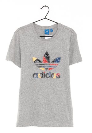 Grå Adidas t-shirt med korte ærmer, flerfarvet broderet logo og tekst, hængende på en sort plastikbøjle mod hvid baggrund.