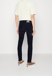 Persona vistiendo jeans ajustados negros, una camiseta blanca de manga corta y zapatillas altas marrones, de pie frente a un fondo blanco liso.