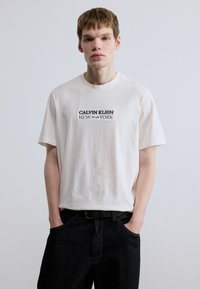 Λευκό βαμβακερό t-shirt με στρογγυλή λαιμόκοψη και κοντά μανίκια, με μαύρη εκτυπωμένη επιγραφή "CALVIN KLEIN NEW EST. 1968 YORK." Συνδυασμένο με σκούρο τζιν παντελόνι.