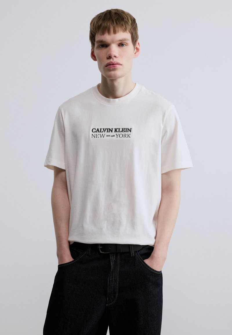 Λευκό βαμβακερό t-shirt με στρογγυλή λαιμόκοψη και κοντά μανίκια, με μαύρη εκτυπωμένη επιγραφή "CALVIN KLEIN NEW EST. 1968 YORK." Συνδυασμένο με σκούρο τζιν παντελόνι.