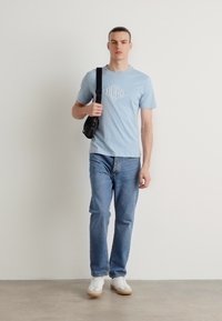 Lyseblå t-skjorte med kort erme og grafisk design, blå jeans og hvite sneakers med gummisåler; svart crossover-veske på skulderen.