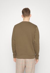 Felpa marrone in pile con collo rotondo, polsini e orlo a costine. Dettagli di cucitura visibili, indossata sopra pantaloni a check.