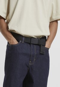Črni tkani pas z pravokotno zaponko, nosi se čez temne jeans hlače. Srajca je svetlo bež. Vidna šivanka na jeansu.