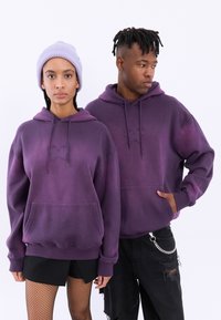 Lilla hettegensere laget av mykt stoff har en frontlomme og snorer i hetta. En subtil logo er brodert på brystet.