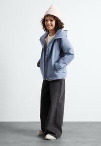 Casaco isolado azul claro com capuz, fecho frontal e bolsos laterais, combinado com calças largas pretas e um gorro de malha rosa.