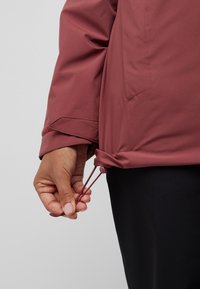 Veste bordeaux avec une surface lisse, dotée de cordons ajustables à l'ourlet et d'un poignet ajusté. L'arrière-plan est un gris uni.
