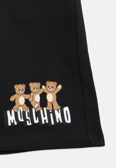 Τρεις καφέ αρκουδάκια στέκονται πάνω από το λευκό λογότυπο "MOSCHINO" σε μαύρο ύφασμα, πιθανόν μέρος ενός ρούχου.