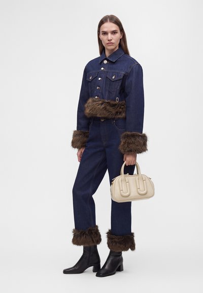 Femme portant une veste et un pantalon en denim bleu foncé avec une bordure en fourrure marron aux poignets et à la taille, tenant un petit sac à main beige et portant des bottes noires.