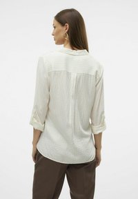 Blouse blanche avec des rayures verticales subtiles et des détails complexes, manches retroussées et coupe décontractée. Vue de dos affichée.