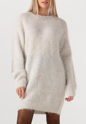 Donna che indossa un abito lungo e ampio con maniche lunghe, in maglia morbida di colore bianco sporco, in piedi davanti a uno sfondo bianco semplice.