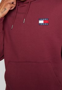 Bordowy sweatshirt z kapturem z przednią kieszenią, z prostokątnym haftowanym logo w kolorach niebieskim, czerwonym i białym z napisem "TOMMY JEANS."