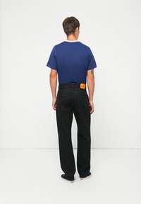 Pantalones de mezclilla negros con bolsillos traseros, combinados con una camiseta azul. Etiqueta amarilla notable en la cintura, con un diseño simple y corte recto.