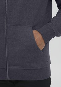 Tamsiai pilkas zip-up gobtuvė su tekstūruotu audiniu, raukšlėtais rankogaliais ir kengūros kišeniu, su akcento logotipu šalia apvado.