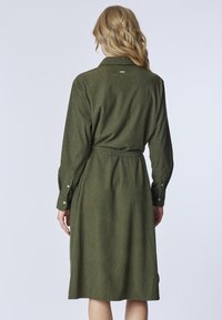 Abito verde oliva a maniche lunghe con colletto da camicia e vita con cintura. Tessuto strutturato e polsini con bottoni. Design di lunghezza media.