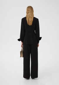 Pantaloni larghi neri abbinati a un blazer su misura dello stesso colore. Il tessuto è liscio con una leggera lucentezza, con polsini bottonati e una borsa con stampa leopardata.