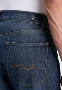 Mörkblå denimjeans med en distinkt bakficka som har ett vågigt brunt sömmönster och en liten tygetikett nära midjan.