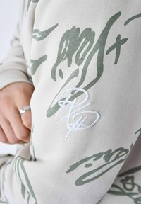 Lys grå sweatshirt med grønne abstrakte mønstre og hvid broderet logo. Stoffet ser blødt ud med en glat tekstur.