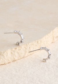 Silberne Creolen mit rechteckigen und runden klaren Steinen, geschwungenem Design und geraden Stiften auf einer strukturierten Oberfläche.