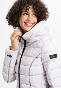 Lichtgrijze pufferjack met een hoge kraag, ritssluiting en zwarte accenten. Bevat gestikte panelen en een logo-embleem op de mouw.