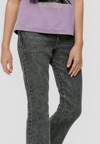 Kind met een zwart vervaagde spijkerbroek en een lavendel Batgirl T-shirt, staande met de handen ontspannen langs hun zij.