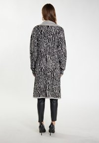 Langer, gemusterter Cardigan in Schwarz und Grau mit einem Schalkragen. Das Design zeichnet sich durch eine strukturierte Optik aus. Wird über enganliegenden schwarzen Hosen und High Heels getragen.