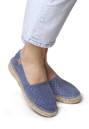 Blue suede espadrilles with decorative metal studs and woven jute soles; ankle-length denim jeans are visible, cuffed at the hem.