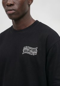 Chemise noire à manches longues portée par une personne, avec le texte blanc déformé "DROPSIZE" imprimé sur la partie supérieure gauche de la poitrine.