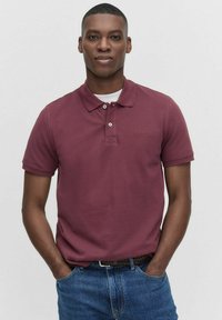 Mörkröd polo skjorta i bomull, med krage, tvåknappsstängning och subtilt bröstlogo. Kombinerad med blå jeans.