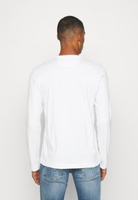 Biała koszulka z długim rękawem, wykonana z gładkiej bawełny, z okrągłym dekoltem, widocznym szyciem i luźnym krojem. Noszona z niebieskimi dżinsami.