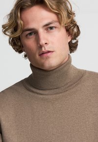 Maglione a collo alto marrone realizzato in tessuto morbido, caratterizzato da una superficie liscia e dettagli a coste nel colletto, progettato per una vestibilità aderente.