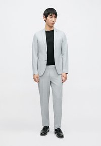 Hellgrauer Anzug mit einreihigem Blazer und passenden Hosen. Darüber wird ein schwarzes, glatt strukturiertes T-Shirt getragen, ergänzt durch schwarze Schuhe.