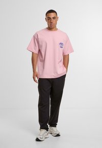 Pink cotton t-shirt with short sleeves, featuring blue text on the chest. Paired with black pants and light sneakers, standing against a white background.