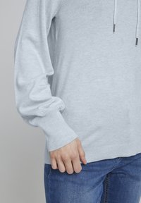 Jasnoniebieski sweter z prążkowanymi mankietami i dołem, o luźnym kroju, z sznurkiem przy dekolcie. Noszony na dżinsach.