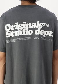 Camiseta de algodón gris con un gran gráfico blanco en la parte posterior que incluye el texto "Originals Studio dept." y un pequeño logotipo, corte relajado.