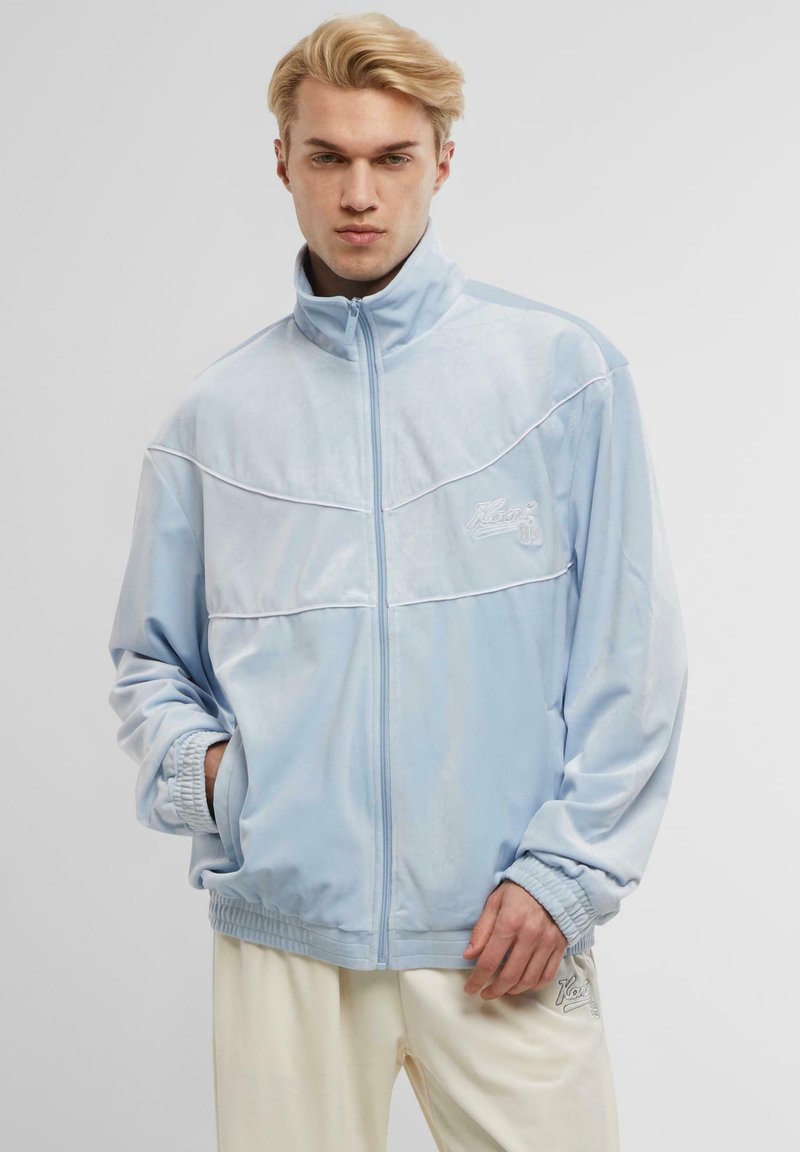 Himmelblaues Veloursjackett mit hohem Kragen, elastischen Bündchen und Taillenbund. Verziert mit weißem Besatz und Logo auf der linken Brust.